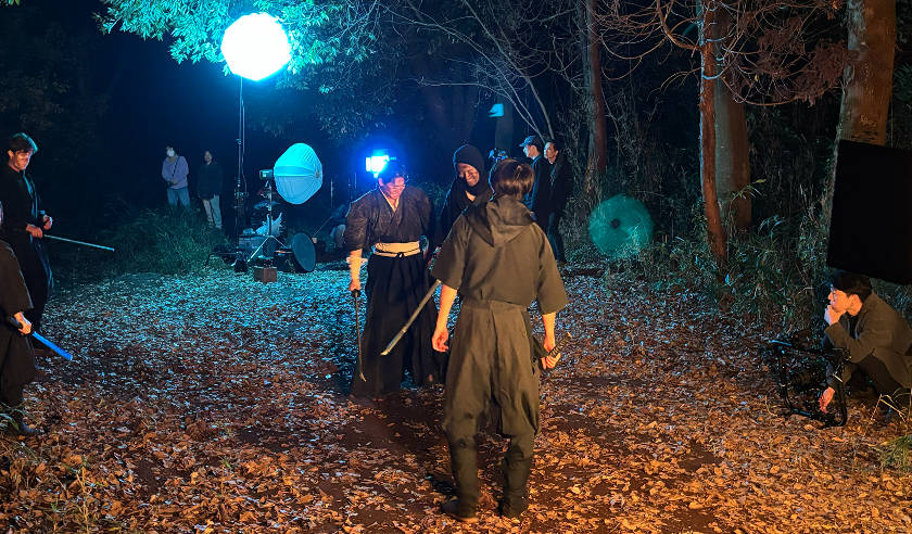 夜の森の中で、着物や忍者のような衣装をまとった演者たちが殺陣（たて）の撮影を行っているシーンです。